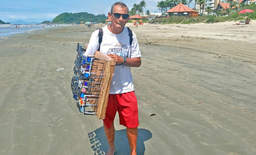 Jonas caminha e mostra os óculos de sol aos turistas, todos os dias, na praia do centro, em Itanhaém