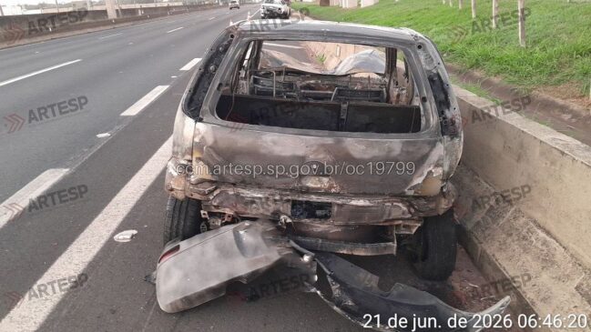 Carro carbonizado aparece parado no acostamento de uma rodovia, ao lado de uma barreira de concreto