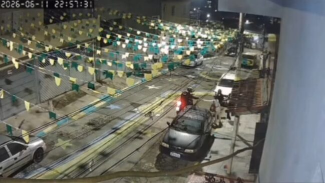 Imagem noturna de câmera de segurança mostrando uma rua residencial enfeitada com bandeirinhas de festa junina verdes e amarelas. No centro, dois homens em uma motocicleta abordam um pedestre na calçada, ao lado de um carro escuro estacionado. No canto superior esquerdo, há um indicador de data e hora digital