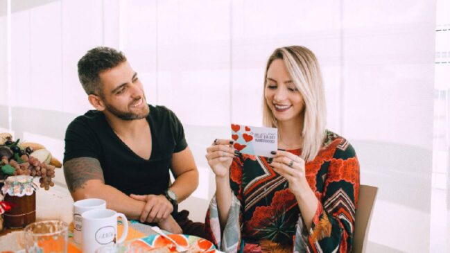 Casal formado por homem branco, de cabelos escuros, e mulher branca, loira, em mesa de jantar, ela está sorrindo lendo um cartão de Dia dos Namorados