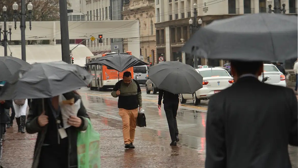 Pessoas caminham sob chuva em uma rua de São Paulo, segurando guarda-chuvas pretos. Em destaque, um homem de calça caramelo e casaco escuro anda pelo asfalto molhado. Ao fundo, vê-se o trânsito com um ônibus laranja, carros brancos e edifícios urbanos.