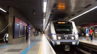 Um trem moderno do metrô, nas cores prata e azul com a identificação H55 na frente, está parado nos trilhos da Estação República. À esquerda, a plataforma está vazia e exibe uma placa vermelha na parede com o nome "República". À direita, um homem de camiseta azul e uma mulher de blusa vermelha aguardam na plataforma oposta, próximos a uma escada rolante