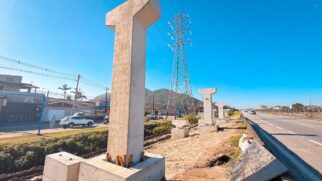 Fileira de grandes pilares de concreto pré-moldado em formato de "T" erguidos em um canteiro de obras de terra batida na lateral de uma rodovia