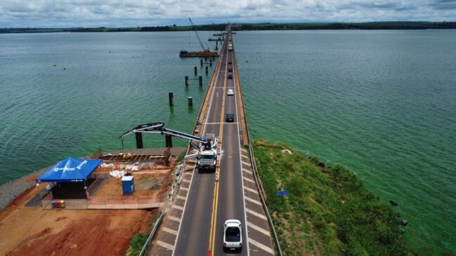 Vista aérea de uma ponte sobre um lago com tráfego de veículos em ambos os sentidos. Ao lado da estrutura, máquinas e equipes trabalham em obras de ampliação ou construção de uma nova travessia, com pilares já instalados na água. A imagem mostra a extensão da ponte e o avanço das intervenções de infraestrutura na região