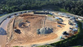 Vista aérea de um canteiro de obras em uma área de terraplanagem cercada por mata densa. No local, há tratores, caminhões de carga, montes de terra e brita, além de estruturas de apoio e contêineres. Uma pista asfáltica em curva contorna o canteiro, com veículos estacionados na lateral