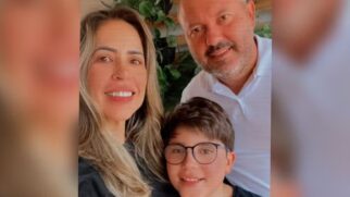 Foto no estilo selfie mostrando uma mulher sorridente em primeiro plano à esquerda, um homem de camisa polo branca logo atrás à direita, e um menino de óculos posicionando-se à frente, entre os dois. Ao fundo, nota-se uma parede com plantas decorativas