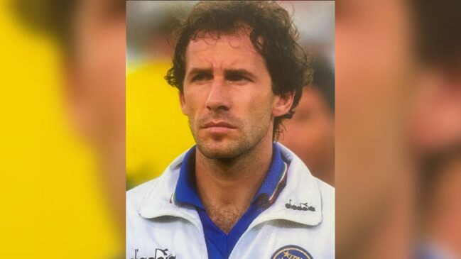 Retrato em plano médio fechado do ex-zagueiro italiano Franco Baresi em seus tempos de jogador. Ele é um homem de pele clara, cabelos cacheados castanhos e olhar sério fixado à frente. Veste uma jaqueta esportiva branca de treino da Seleção Italiana, da marca Diadora, com gola dobrada azul e detalhe em verde e vermelho nas bordas da gola. No peito esquerdo, aparece parte do escudo da Federação Italiana de Futebol (FIGC). O fundo da imagem está desfocado em tons quentes de amarelo e alaranjado.