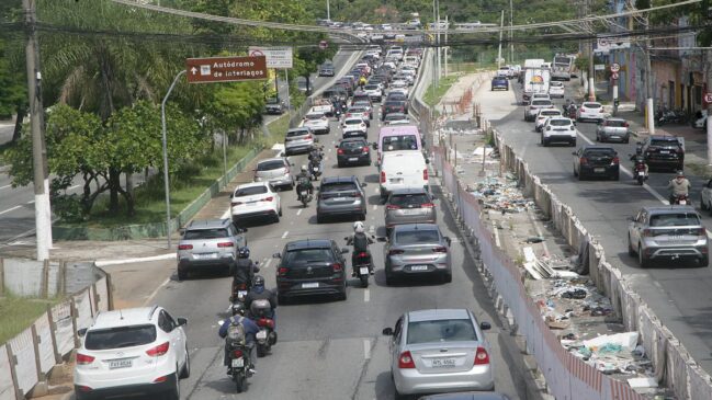 Fotografia diurna e elevada de uma avenida movimentada com tráfego intenso de carros, peruas e motocicletas. À esquerda, destaca-se uma placa turística marrom indicando a direção em frente para o "Autódromo de Interlagos". Ao centro e à direita, a pista é margeada por um trecho em obras delimitado por tapumes brancos e barreiras de concreto, com entulho acumulado no canteiro. Ao fundo, o trânsito segue adensado sobre um trecho de elevação sob fiação aérea.