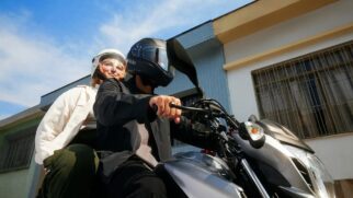 Fotografia diurna em ângulo inferior de um homem pilotando uma motocicleta cinza com uma passageira na garupa. O condutor veste jaqueta escura e capacete preto fechado que exibe o logotipo "Uber Moto" escrito em letras brancas na lateral