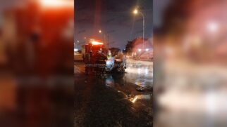 Foto noturna de um acidente viário em uma rodovia com asfalto molhado e reflexos de luzes. No centro, bombeiros com uniformes escuros e faixas refletivas prestam socorro ao redor de um veículo de passeio severamente destruído e amassado