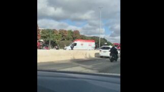 A foto mostra as pistas da Marginal Tietê separadas por uma barreira de concreto. Na pista do sentido oposto, ocorre o atendimento a um acidente. Uma ambulância branca e vermelha do SAMU (192) está parada no local, acompanhada de socorristas, motociclistas passando e um carro branco de passeio. Ao fundo, há árvores verdes sob um céu azul parcialmente nublado.