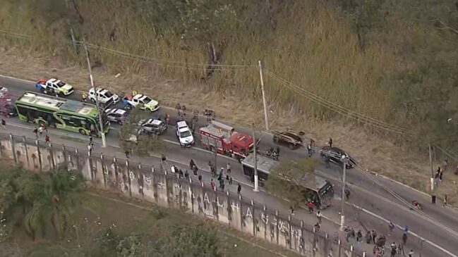 Fotografia aérea em ângulo panorâmico mostrando uma operação de resgate após acidente de trânsito em uma via da Zona Norte de São Paulo. Na pista, veem-se dois ônibus do transporte público municipal (um verde em destaque no canto esquerdo), um caminhão vermelho do Corpo de Bombeiros, viaturas da Polícia Militar e carros de passeio retidos.