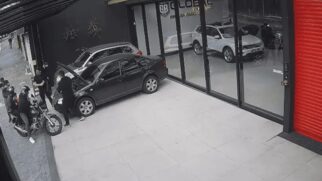 Captura de tela em ângulo elevado de uma câmera de segurança mostrando a fachada da loja "GB Global Veículos". No pátio externo de piso claro, um carro preto está estacionado com o capô aberto e um homem de preto mexe no motor. No canto esquerdo, dois homens usando capacetes estão montados em uma motocicleta preta parada, virados em direção ao grupo