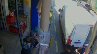 Frame de câmera de segurança em ângulo superior mostrando a calçada em frente a um mercado. No centro, três homens lutam corporalmente; um deles de boné verde e outro de camisa preta. Um revólver prateado está no chão entre os indivíduos em luta. Um furgão branco Fiat Ducato está estacionado à direita com uma placa brasileira visível
