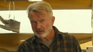 Fotografia em primeiro plano (close-up) do ator Sam Neill. Ele é um homem branco de idade avançada, com cabelos grisalhos curtos e barba e bigode também grisalhos. Ele veste uma camisa xadrez em tons escuros de verde e marrom sobre uma camiseta cinza. Sua expressão é séria e atenta, olhando ligeiramente para o lado