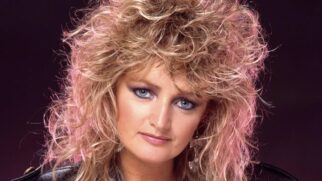 Retrato em plano fechado (close-up) da cantora Bonnie Tyler nos anos 1980. Ela tem cabelos loiros volumosos, repicados e cacheados, típicos da época, com reflexos de iluminação arroxeada nas pontas.
