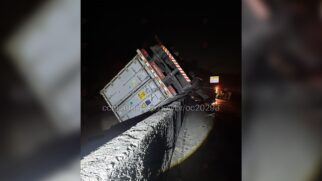 Fotografia noturna do tombamento de uma carreta com contêiner prata sobre a mureta de concreto (barreira New Jersey) de uma rodovia. O veículo de carga aparece inclinado e equilibrado sobre a divisória de pistas, expondo o chassi traseiro e a estrutura do contêiner.