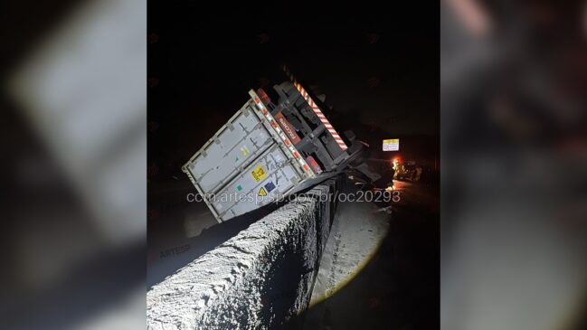 Fotografia noturna do tombamento de uma carreta com contêiner prata sobre a mureta de concreto (barreira New Jersey) de uma rodovia. O veículo de carga aparece inclinado e equilibrado sobre a divisória de pistas, expondo o chassi traseiro e a estrutura do contêiner.