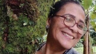 Fotografia em primeiro plano (close-up) de uma mulher de meia-idade sorrindo. Ela tem cabelos escuros presos, usa óculos de grau com armação avermelhada e brincos prateados. A mulher está com o rosto recostado no tronco de uma árvore densamente coberto por musgo verde