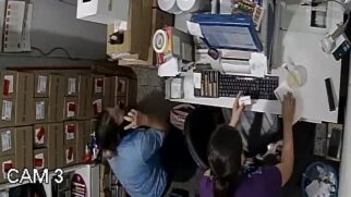 Imagem de ângulo superior capturada por uma câmera de segurança interna identificada como "CAM 3" no canto inferior esquerdo. Em um escritório ou balcão de atendimento, uma mulher de blusa roxa está sentada de costas em frente a um computador, segurando um pequeno papel branco retangular. Ao lado dela, à esquerda, outra funcionária com uniforme azul manipula caixas de papelão empilhadas. A mesa de trabalho exibe um teclado, organizadores, bobinas de papel e um monitor com o reflexo de telas azuis de sistema
