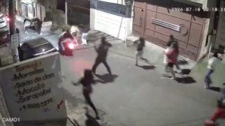 Captura de tela em ângulo superior de uma câmera de segurança noturna identificada como "CAM01" no canto inferior esquerdo. Em uma rua residencial iluminada por postes, várias pessoas correm em direções opostas em sinal de desespero e correria. No centro, destaca-se um homem de costas correndo, e ao fundo uma motocicleta em movimento com a lanterna traseira acesa