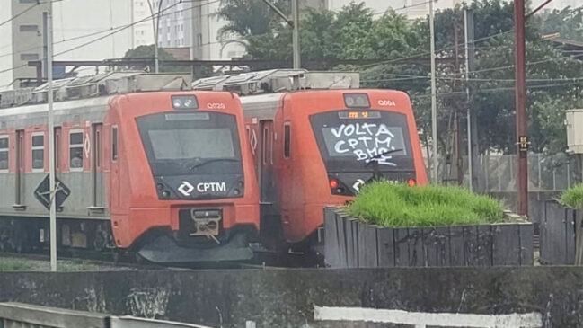 Fotografia diurna de dois trens da CPTM nas cores vermelha e cinza estacionados lado a lado em um pátio sobre os trilhos. O trem da esquerda exibe a identificação Q020 e o logotipo da CPTM na frente. O trem da direita, com identificação Q004, traz uma pichação em tinta branca no para-brisa frontal com a mensagem "VOLTA CPTM". Em primeiro plano, vê-se uma estrutura de concreto com vegetação e, ao fundo, fiação aérea sob céu nublado