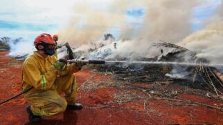 Agente do Corpo de Bombeiros agachado sobre solo de terra vermelha, vestindo uniforme antichamas amarelo, capacete laranja, óculos de proteção e máscara respiratória azul. Ele segura firmemente o esguicho de uma mangueira e dispara um jato de água direto contra uma grande pilha de galhos e vegetação seca em chamas. O fogo gera uma densa nuvem de fumaça branca e cinza que toma conta do cenário.