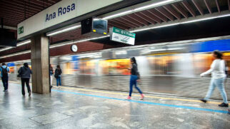 Vista em perspectiva da plataforma subterrânea da Estação Ana Rosa do Metrô. À esquerda, pedestres caminham pela plataforma de piso cinza escuro polido, delimitada por uma faixa amarela de segurança e uma linha azul no chão. No topo esquerdo, destaca-se uma grande placa de sinalização branca com a inscrição "Ana Rosa" em azul. Um trem de aço escovado passa pelas vias em alta velocidade à direita, gerando um efeito de borrão de movimento (motion blur). Placas aéreas indicam o destino da Linha 2-Verde em direção à Vila Prudente.