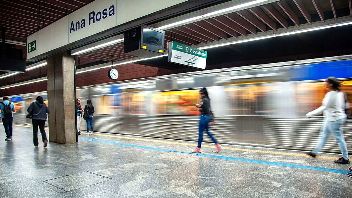 Vista em perspectiva da plataforma subterrânea da Estação Ana Rosa do Metrô. À esquerda, pedestres caminham pela plataforma de piso cinza escuro polido, delimitada por uma faixa amarela de segurança e uma linha azul no chão. No topo esquerdo, destaca-se uma grande placa de sinalização branca com a inscrição "Ana Rosa" em azul. Um trem de aço escovado passa pelas vias em alta velocidade à direita, gerando um efeito de borrão de movimento (motion blur). Placas aéreas indicam o destino da Linha 2-Verde em direção à Vila Prudente.
