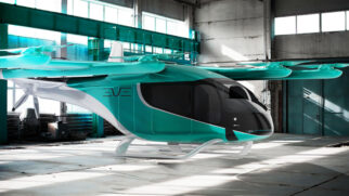 Renderização digital de uma aeronave futurista de pouso e decolagem vertical (eVTOL) nas cores verde-turquesa, branca e preta, estacionada dentro de um grande galpão industrial com piso de concreto e janelas altas.