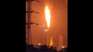 Fotografia noturna mostrando uma labareda alta e intensa de fogo saindo da torre de queimador de segurança (flare) de um polo petroquímico, iluminando o céu em tom alaranjado.