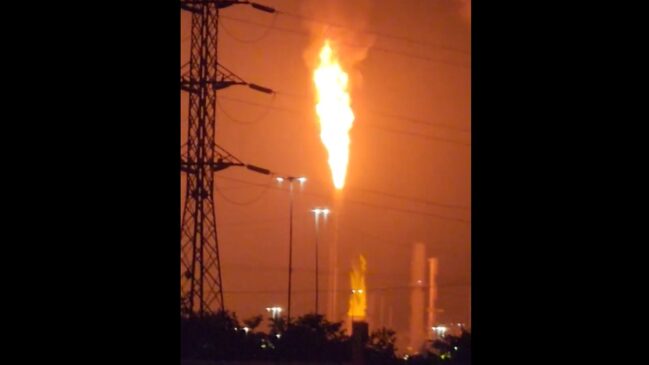 Fotografia noturna mostrando uma labareda alta e intensa de fogo saindo da torre de queimador de segurança (flare) de um polo petroquímico, iluminando o céu em tom alaranjado.