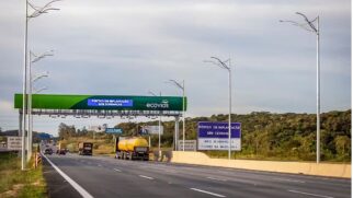 Fotografia diurna de uma rodovia larga com várias faixas de rolamento. Acima da pista, cruza um grande pórtico metálico verde com o logotipo da Ecovias e um painel digital azul que exibe a mensagem "Pórtico em implantação sem cobranças". No acostamento à direita, uma placa retangular reforça o aviso informando "Pórtico em implantação sem cobrança - Mais segurança e fluidez na Imigrantes".