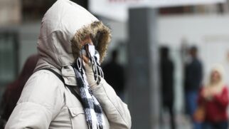Pedestre fotografada de perfil usando um casaco de inverno grosso e claro com o capuz forrado de pelos cobrindo a cabeça. Ela usa luvas e leva a mão ao rosto para se proteger do vento, cobrindo o nariz e a boca com um cachecol xadrez nas cores preto e branco