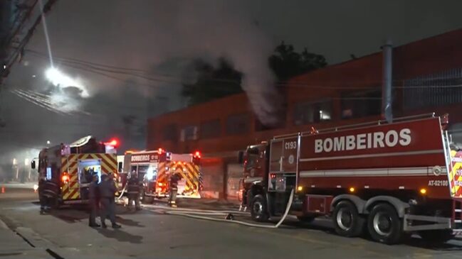 Fotografia noturna mostrando a operação do Corpo de Bombeiros durante o combate a um incêndio em prédio comercial de fachada vermelha no Bom Retiro, em São Paulo. No lado direito, em destaque, há um grande caminhão-tanque vermelho com a palavra "BOMBEIROS" escrita em letras brancas garrafais. Ao centro e à esquerda, veem-se outros veículos de emergência com sinalizadores acesos, mangueiras estendidas pelo asfalto e bombeiros fardados na pista. Fumaça cinza-escura sai da parte superior do imóvel.
