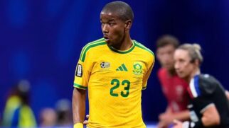 Fotografia de cintura para cima de um jogador de futebol negro em campo durante uma partida. Ele veste a camisa amarela da seleção da África do Sul, que possui detalhes e listras nos ombros em verde, além do número 23 centralizado no peito. O atleta tem o cabelo bem curto, expressão concentrada e olha para o lado esquerdo