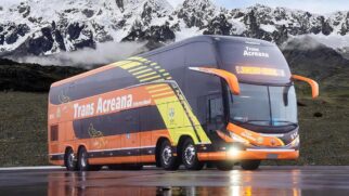 Ônibus rodoviário de dois andares (double-decker) da empresa Trans Acreana estacionado sobre uma pista molhada que reflete sua imagem. O veículo possui pintura predominantemente laranja, com a base preta e grafismos geométricos em amarelo