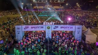 Fotografia aérea noturna em ângulo elevado mostrando a largada da corrida de rua SP City Marathon na Praça Charles Miller. Em primeiro plano, milhares de corredores preenchem a avenida e cruzam a linha de partida sob um grande pórtico iluminado com as inscrições "START" e "RUN SPCITY '26 MARATHON".