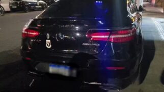Fotografia noturna em plano fechado da traseira de um SUV cupê de luxo preto, da marca Mercedes-AMG. O carro apresenta a estrela de três pontas da Mercedes-Benz no centro, a inscrição cromada "AMG" no lado direito e um pequeno adesivo em formato de brasão de segurança ou corporativo no lado esquerdo da tampa do porta-malas
