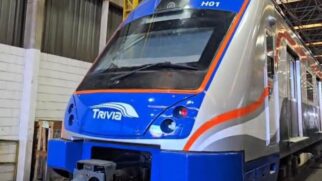 Fotografia em ângulo de três quartos da cabine frontal de um novo trem de passageiros moderno, estacionado no interior de uma oficina ou galpão industrial. A máscara frontal do veículo é azul brilhante, com a logomarca "TRIVIA" estampada em branco no centro e a identificação "H01" gravada no topo, acima do para-brisa.