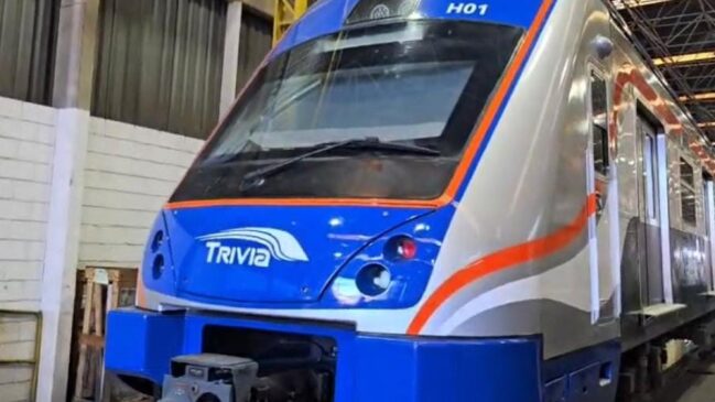 Fotografia em ângulo de três quartos da cabine frontal de um novo trem de passageiros moderno, estacionado no interior de uma oficina ou galpão industrial. A máscara frontal do veículo é azul brilhante, com a logomarca "TRIVIA" estampada em branco no centro e a identificação "H01" gravada no topo, acima do para-brisa.
