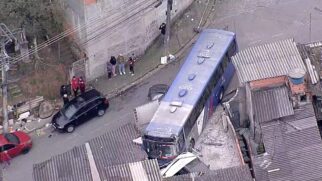 Fotografia aérea tirada de ângulo elevado mostrando um ônibus de transporte público azul e cinza acidentado em uma rua residencial estreita e íngreme. O veículo colidiu de frente contra o muro de uma casa, destruindo a estrutura de concreto e invadindo parte do terreno. Os escombros do muro aparecem espalhados ao redor da parte frontal do ônibus.