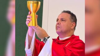 Fotografia de plano médio de um padre homem de pele clara e cabelos escuros, visto de perfil de três quartos. Ele segura um grande cálice dourado reluzente com as duas mãos, elevando-o durante um rito religioso. O sacerdote veste uma casula litúrgica vermelha com acabamento dourado na gola sobre uma túnica alva branca com detalhes bordados nas mangas. No punho esquerdo, ele usa um relógio de pulso escuro. Sua expressão é solene e compenetrada, com o olhar direcionado para cima. Ao fundo, vê-se uma parede dividida entre o tom verde e o branco