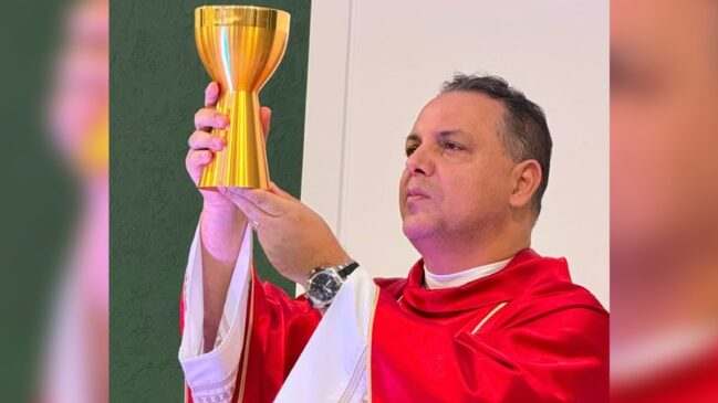 Fotografia de plano médio de um padre homem de pele clara e cabelos escuros, visto de perfil de três quartos. Ele segura um grande cálice dourado reluzente com as duas mãos, elevando-o durante um rito religioso. O sacerdote veste uma casula litúrgica vermelha com acabamento dourado na gola sobre uma túnica alva branca com detalhes bordados nas mangas. No punho esquerdo, ele usa um relógio de pulso escuro. Sua expressão é solene e compenetrada, com o olhar direcionado para cima. Ao fundo, vê-se uma parede dividida entre o tom verde e o branco