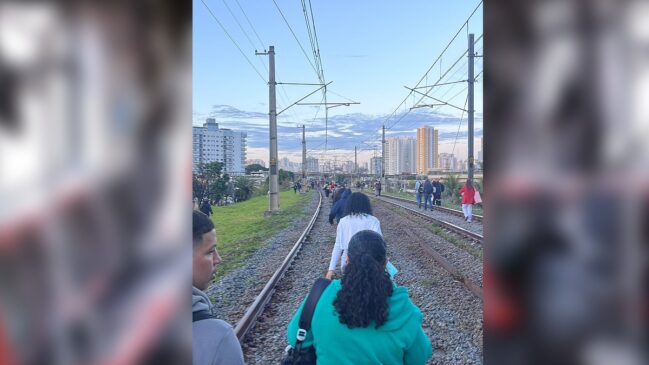 Fotografia vertical em perspectiva traseira de passageiros caminhando pelos trilhos de uma linha de trem. Em primeiro plano, vê-se de costas uma mulher de cabelos escuros e cacheados vestindo um moletom verde-água e bolsa a tiracolo. À sua frente, uma fila de pessoas caminha sobre a brita entre os trilhos de aço. Acima dos trilhos, estendem-se os postes de sustentação e a fiação aérea da rede elétrica