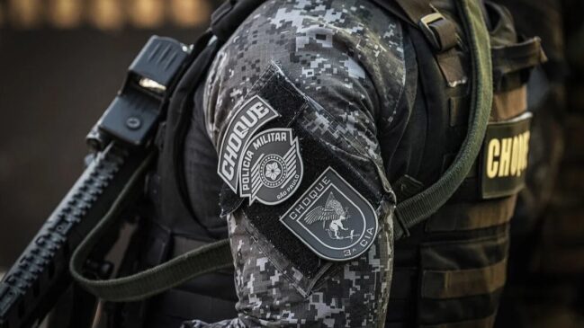 lano fechado no braço e ombro de um policial militar do Batalhão de Choque de São Paulo. O agente veste uma farda tática com padrão de camuflagem digital em tons de cinza e preto. Na manga, destacam-se dois patches (emborrachados) táticos: o superior exibe o brasão estilizado da PMESP com a inscrição "CHOQUE", e o inferior traz o emblema da "3ª CIA" com a figura de um grifo