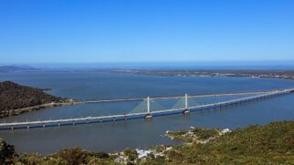 Vista aérea panorâmica em ângulo elevado da Ponte Anita Garibaldi, uma grande estrutura estaiada que cruza uma vasta lagoa de águas escuras. No centro da ponte, destacam-se dois grandes mastros verticais brancos com cabos de sustentação (estais) paralelos dispostos em formato de leque