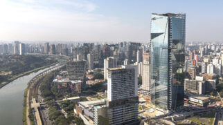 Vista aérea panorâmica e diurna de um trecho de São Paulo ao longo da Marginal Pinheiros. No lado direito, destaca-se um imponente arranha-céu moderno totalmente espelhado, com linhas geométricas marcantes em sua fachada de vidro azulado