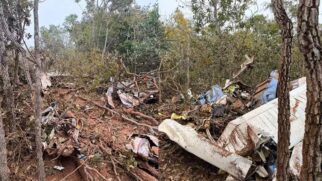 Imagem dividida ao meio mostrando duas perspectivas dos destroços de um acidente aéreo em uma floresta. À esquerda, pedaços de metal retorcido estão espalhados pelo chão de terra vermelha, entre galhos quebrados e árvores. À direita, em plano mais fechado, destaca-se uma grande parte da fuselagem branca e azul da aeronave completamente destruída e amassada, cercada pela vegetação densa