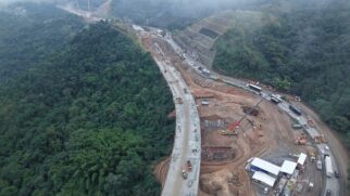 Vista aérea de um amplo canteiro de obras de engenharia na Serra das Araras, na Rodovia Presidente Dutra (BR-116). À esquerda, observa-se uma encosta montanhosa coberta por vegetação nativa sob neblina. Ao centro, destaca-se a construção de uma nova pista e viaduto de concreto, com a presença de guindastes, tratores e caminhões caçamba sobre o solo de terraplanagem. À direita, a pista atual da rodovia segue com tráfego contínuo de caminhões, ônibus e carros de passeio paralelos à área de intervenção